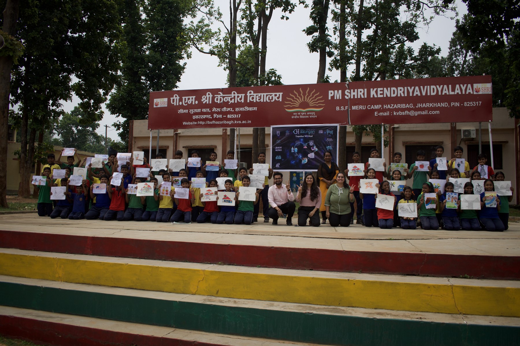 A large school gathering holding drawings in front of the PM SHRI Kendriya Vidyalaya signboard